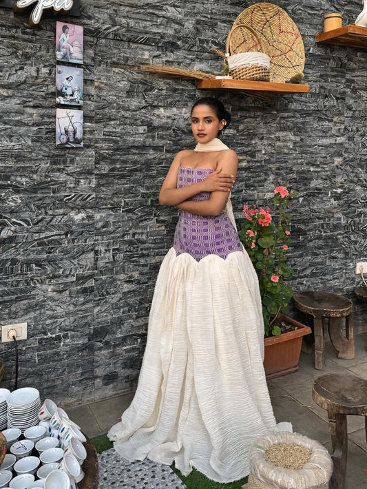 Woman in a purple top and white  Habesha dress standing against a stone wall with decorative items.