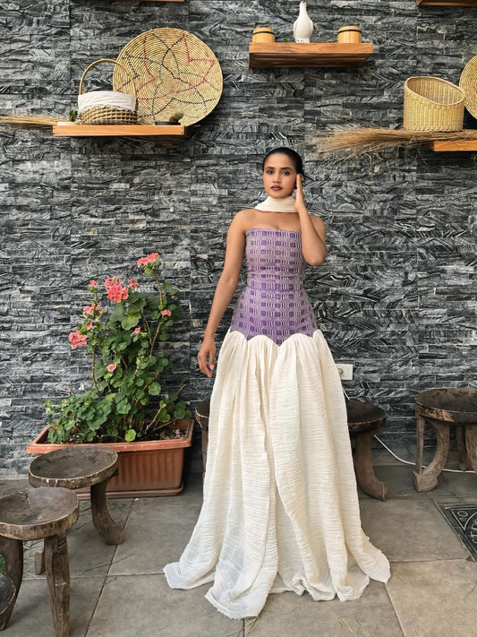 Woman in a purple top and white  Habesha dress standing in front of a stone wall with shelves and plants.