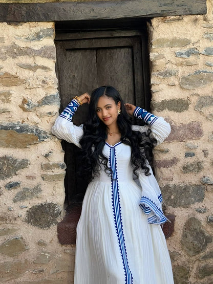 Woman in a white Ethiopian Dress with blue accents standing against a stone wall.
