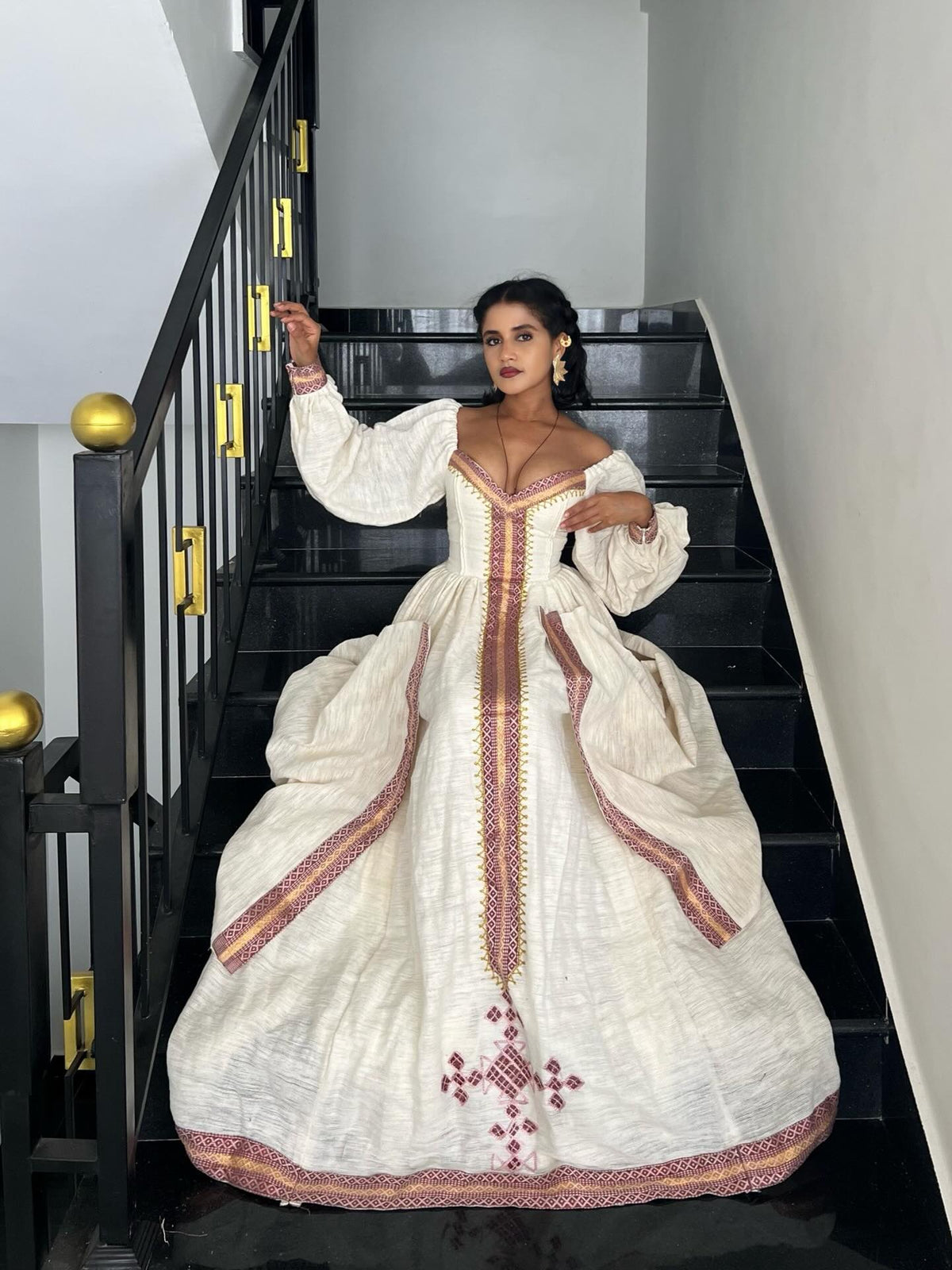 A woman wearing a voluminous off-white medieval-style Ethiopian kemis with red and gold embroidery, posing on a black staircase.