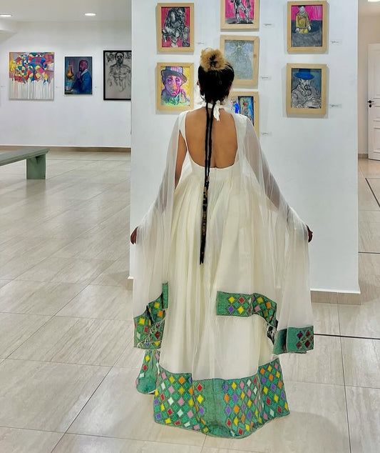 A woman stands with her back to the camera in a white traditional Ethiopian Dress with green patterned borders, in an art gallery with paintings on the walls.