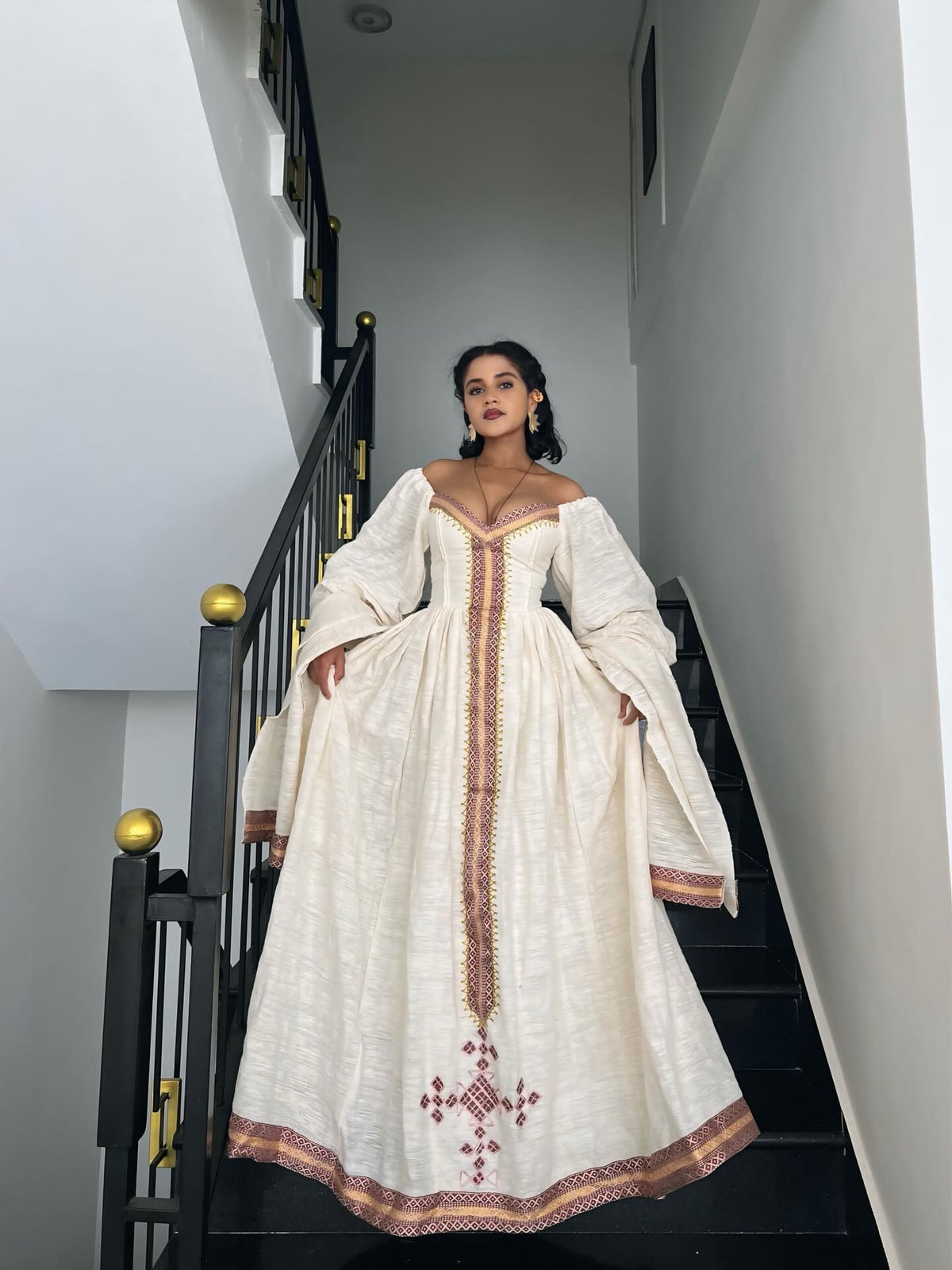 A woman wearing a long off-the-shoulder Ethiopian kemis with red and gold embroidery stands on a black staircase indoors.