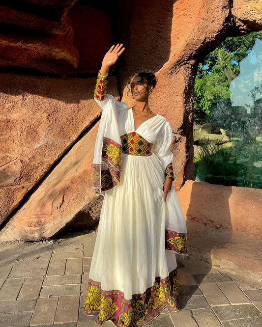 A woman wearing a white maxi Ethiopian Dress with colorful embroidered patterns on the sleeves, waist, and hem, posing outdoors in front of a rustic rock wall.