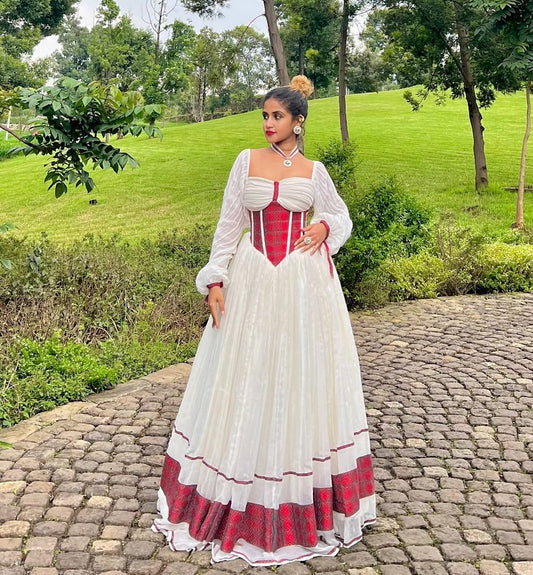 A woman stands outdoors on a cobblestone path wearing a long white Ethiopian Traditional Dress with a red corset-style bodice and red patterned hem, set against a green park background with trees and grass.