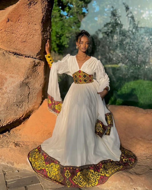 A woman wearing a white maxi Ethiopian Dress with colorful embroidered patterns on the sleeves, waist, and hem, posing outdoors in front of a rustic rock wall.