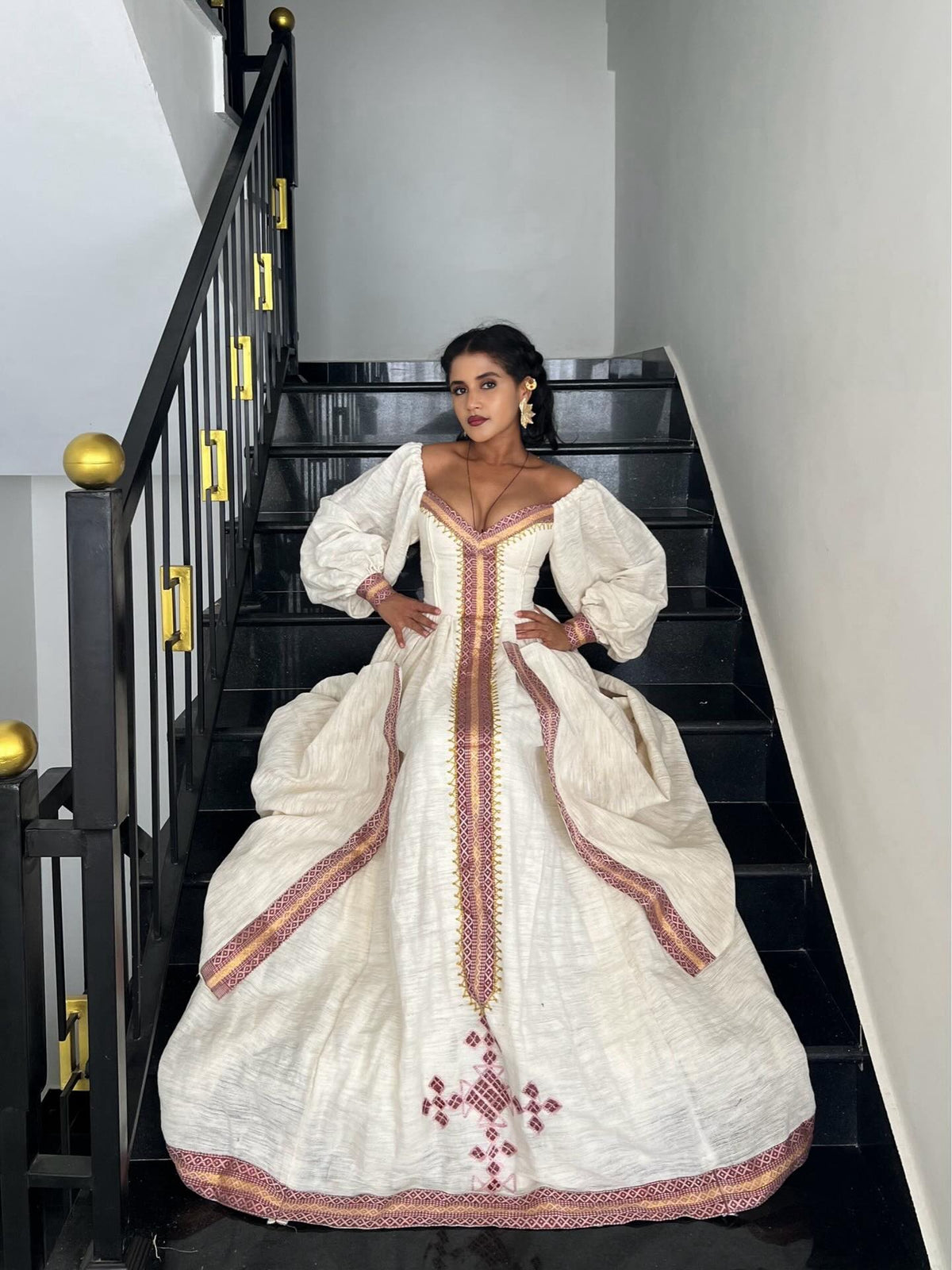 A woman wearing a voluminous off-white Ethiopian kemis with red and gold embroidery, posing on a black staircase.