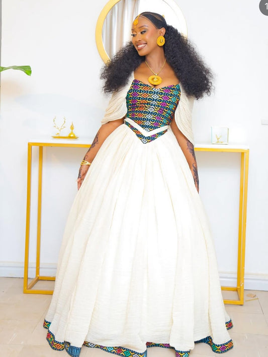 A woman modeling a traditional Habesha Kemis dress with a voluminous white skirt, a colorful embroidered bodice, and a matching draped cape, standing in a brightly lit room with a gold console table.