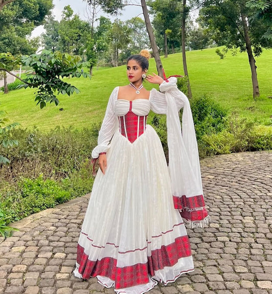 A woman stands outdoors on a cobblestone path wearing a traditional white and red Ethiopian Traditional Dress with a fitted red corset-style bodice, sheer long sleeves, and a full skirt with red patterned trim. Trees and a grassy hill are in the background.