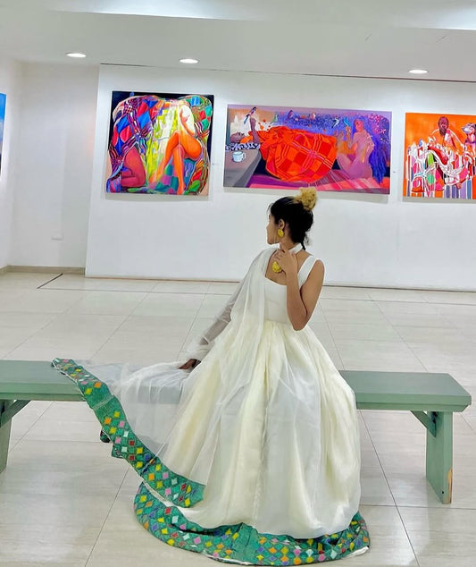 A woman in a white traditional Ethiopian Dress with a green patterned border sits on a bench in a gallery, looking at colorful paintings on the wall.