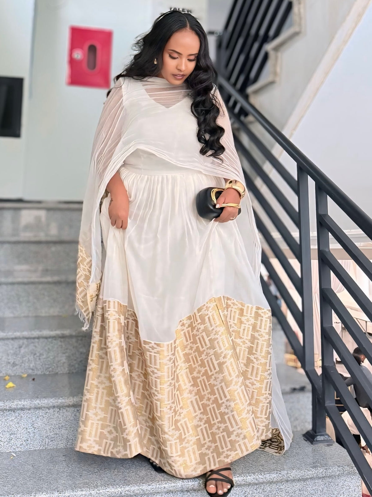 A woman wearing a white and beige patterned traditional ethiopian dress with a sheer white dupatta, standing on outdoor concrete stairs next to a black metal railing.