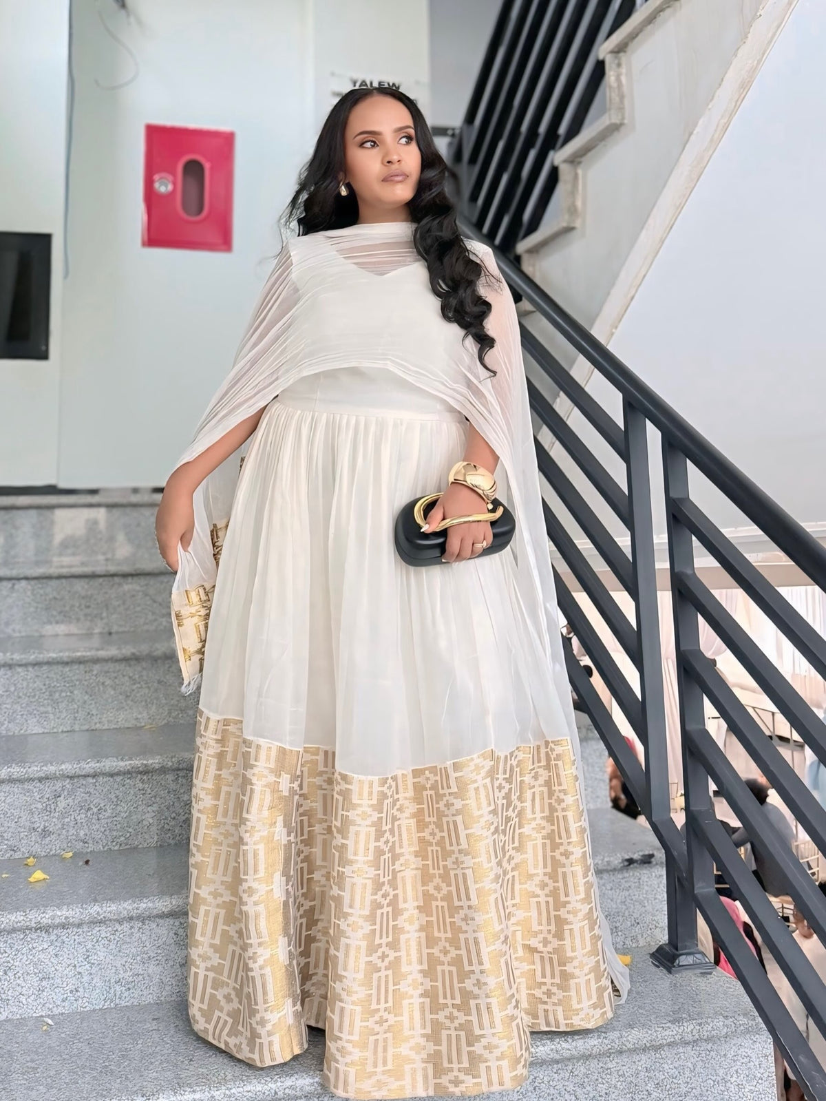 A woman stands on outdoor concrete stairs wearing a traditional ethiopian dress consisting of a white and beige patterned dress with a sheer white dupatta. She is holding a small black clutch bag with gold detailing.