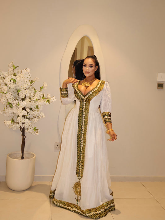 A woman wearing a white traditional long Ethiopian National Dresses with ornate gold embroidery and gold jewelry, posing in front of a white arched mirror in an indoor setting.