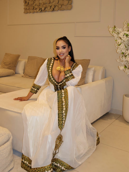 A woman wearing a white traditional long Ethiopian National Dresses with ornate green and gold embroidered trim, seated on a white sofa in a modern living room setting.