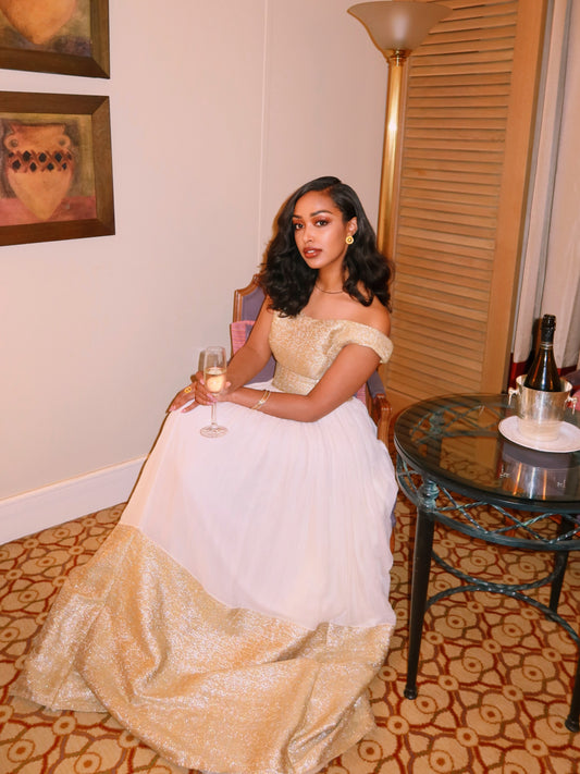 A woman seated indoors wearing a formal off-the-shoulder Ethiopian Dress  with a white tulle upper section and a shimmering gold sequined lower skirt. She holds a glass in a decorated room with framed wall art, a floor lamp, and a small round table.
