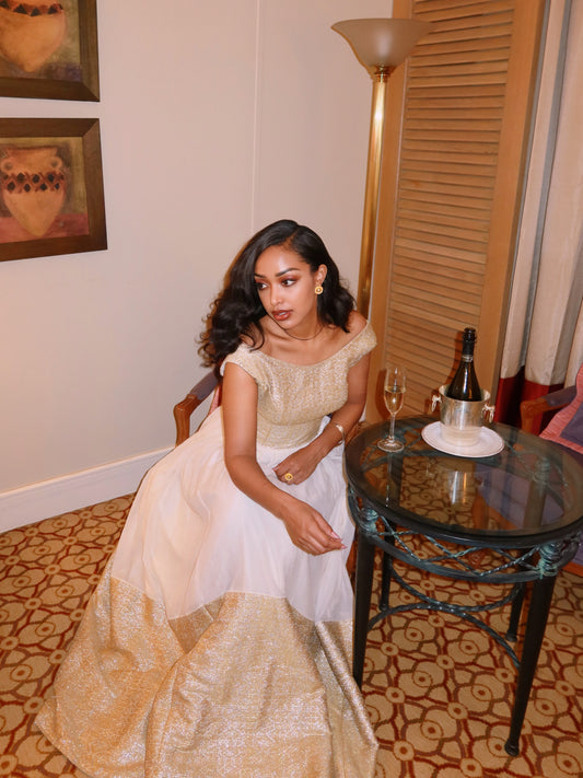 A woman wearing a white and gold off-the-shoulder Ethiopian Dress  sits in a room next to a small glass table with a champagne bottle and glasses.