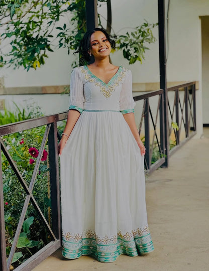 Woman in a light-colored Habesha Wedding Dress with green accents standing on a balcony with greenery.