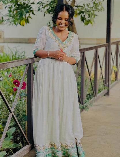 Woman in a long, light-colored habesha dress with green accents standing on a wooden bridge in a garden setting.