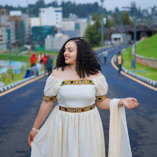 A woman wearing a cream off-the-shoulder Habesha kemis with colorful embroidered borders stands on a paved road in an outdoor urban setting.
