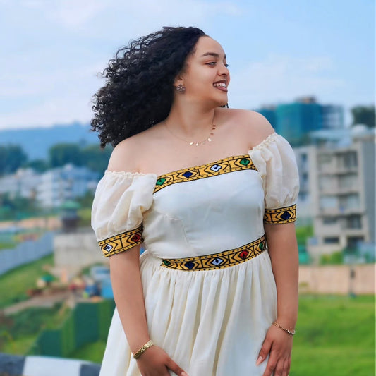 A woman wearing a cream off-the-shoulder Habesha kemis with colorful geometric embroidered trim at the neckline, sleeve cuffs, and waist, standing outdoors with a blurred city background.