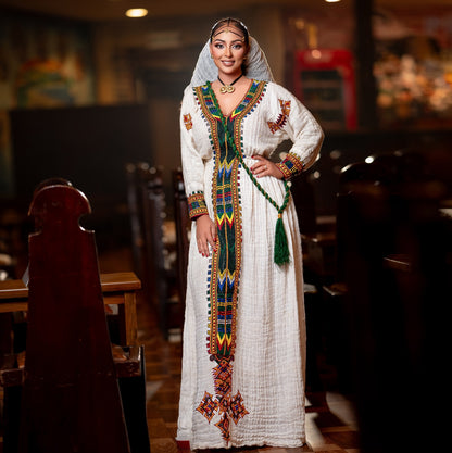 A woman wearing a traditional white embroidered long Ethiopian National Dress with colorful geometric patterns and a matching head covering, standing in a dimly lit indoor setting.