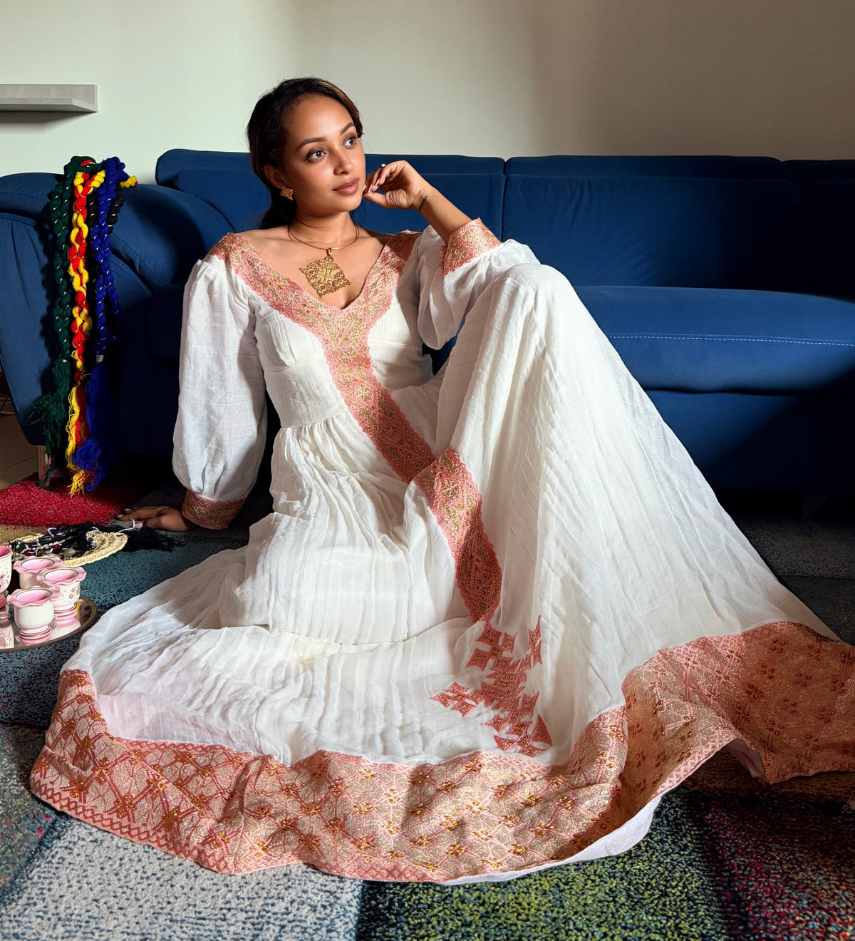 A woman sits on a blue couch wearing a white traditional Ethiopian National Dress with peach-pink embroidered borders and a matching dupatta. The dress has long sleeves, a V-neckline, and detailed embroidery along the neckline and border.