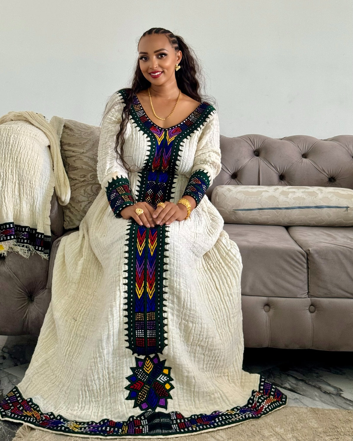 A woman wearing a long white traditional Ethiopian National Dress with colorful geometric embroidery, sitting on a tufted sofa in a living room setting.