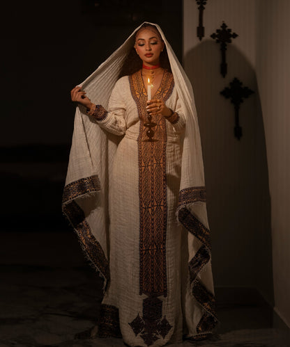 A woman wearing a traditional long white Ethiopian National Dress with ornate brown and black embroidered patterns, holding a lit candle. She has a white head covering draped over her head and stands in a dimly lit interior with dark wall decorations.