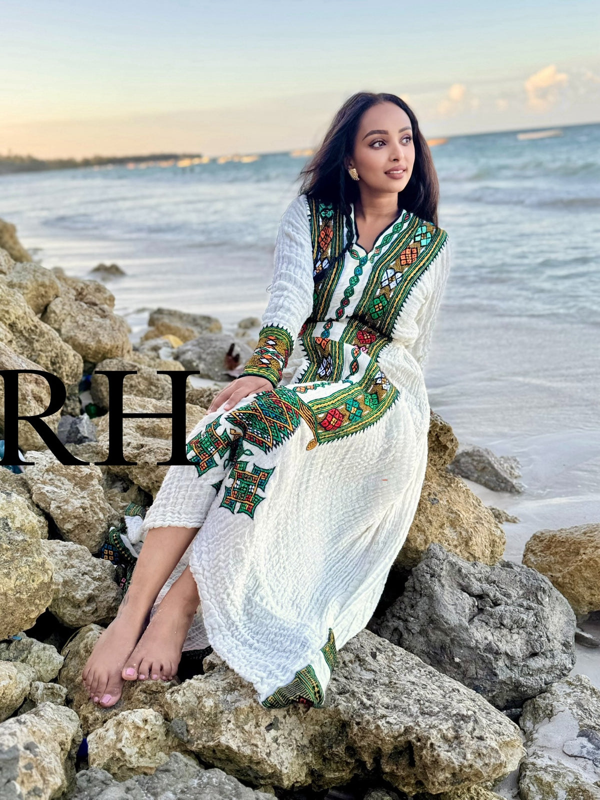 A woman sits on rocks by the ocean wearing a long white embroidered Axum Fetil with green, red, and blue geometric patterns. The brand name 'RH' is overlaid on the left side of the image.