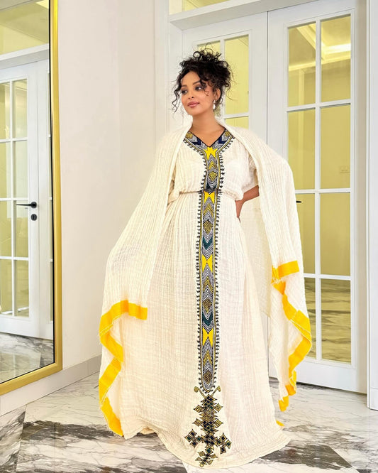 A woman models a traditional Moroccan caftan (Gharama) in cream-colored textured cotton Ethiopian National Dress  with vibrant yellow and blue geometric embroidery and ruffle accents, set against a modern interior background with white glass doors.