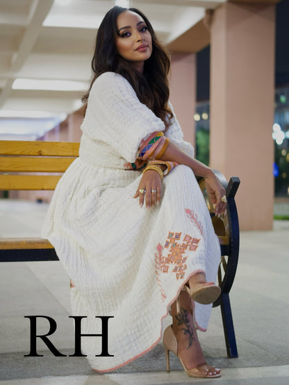 A woman sits on a wooden bench wearing a white textured maxi Ethiopian National Dress with peach embroidery and colorful patterned sleeve cuffs. The image has a large 'RH' watermark in the bottom left corner.