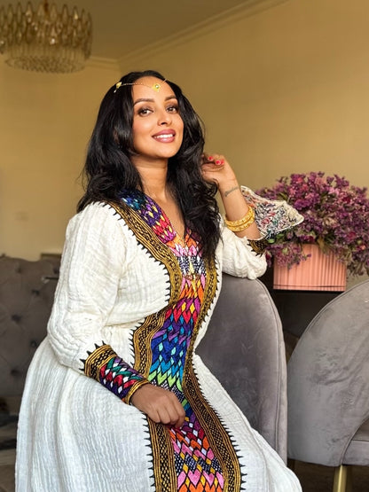 A woman wearing a white traditional embroidered Axum Fetil  with a vibrant multicolor geometric border, posing in an indoor setting with a chaise lounge and flowers in the background.