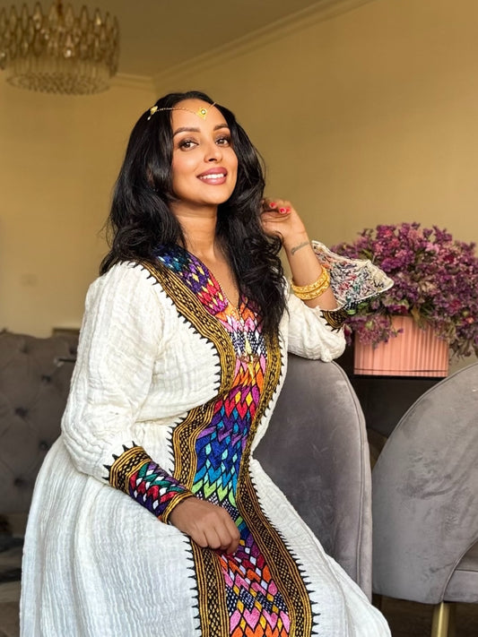 A woman wearing a white traditional embroidered Axum Fetil  with a vibrant multicolor geometric border, posing in an indoor setting with a chaise lounge and flowers in the background.