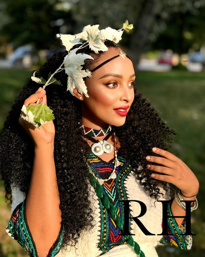 A woman outdoors wearing a white and black patterned top with colorful geometric Ethiopian National Dress, a large silver pendant necklace, bracelets, and a floral headpiece with white flowers. The background is a blurred green park setting, and a large 'RH' watermark is visible in the lower right corner.