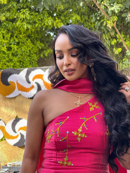 A woman wearing a bright pink one-shoulder Ethiopian National Dress with gold and multicolor embroidered details, posed outdoors in front of green foliage and a patterned chair.