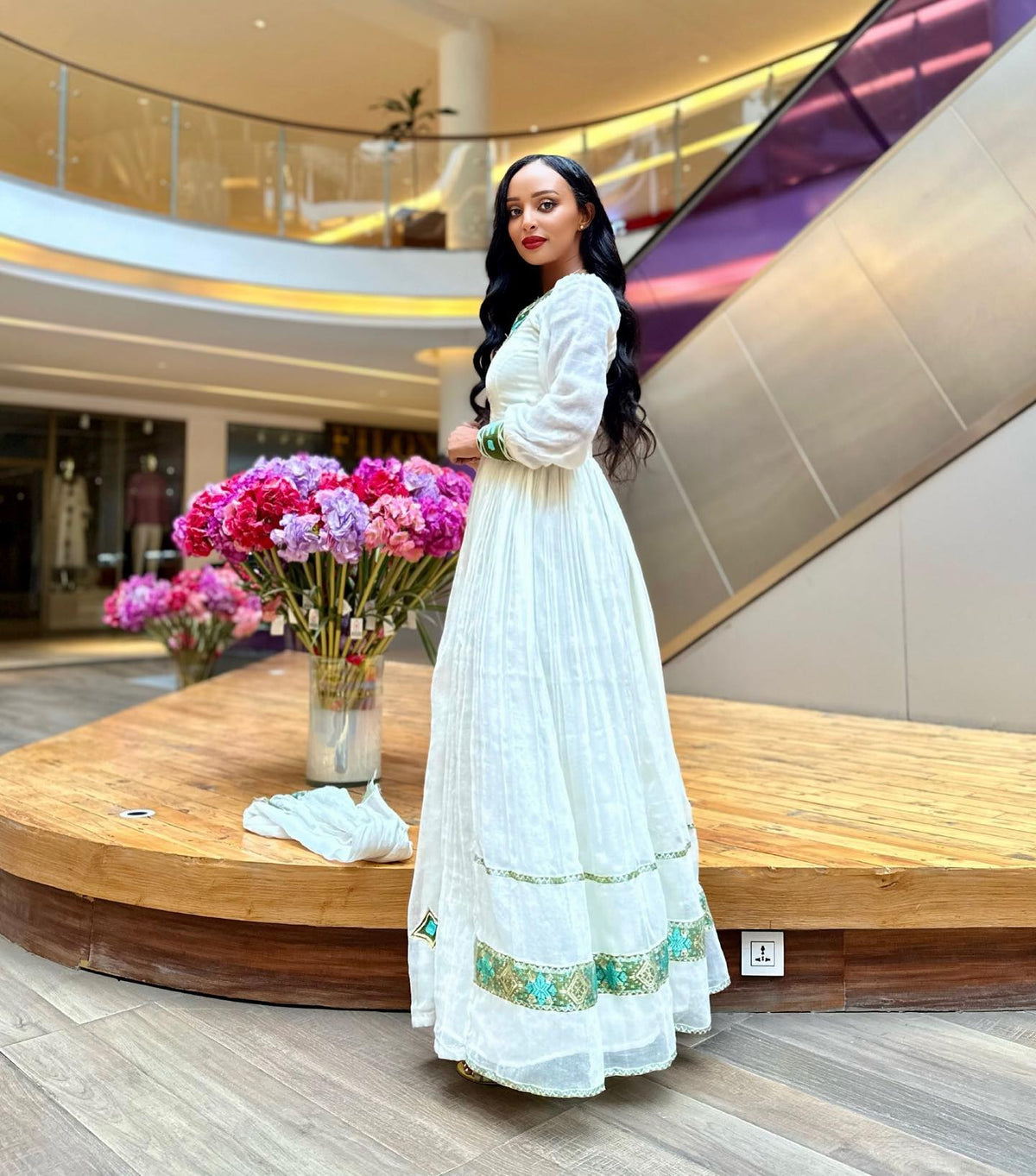 A woman wearing a long white embroidered Ethiopian National Dress stands in a modern indoor mall setting with escalators and flower arrangements in the background.