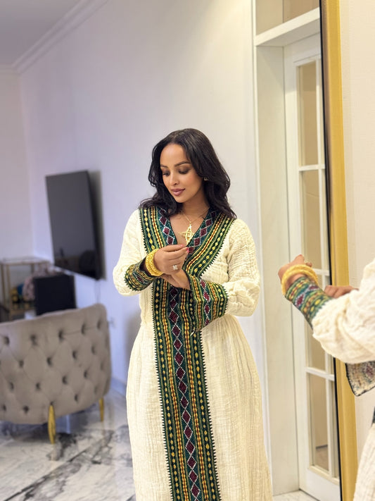 A woman wearing a cream-colored textured Axum Fetil with a prominent green, red, and yellow geometric embroidered border, standing in a modern room with a mirror reflecting her profile.
