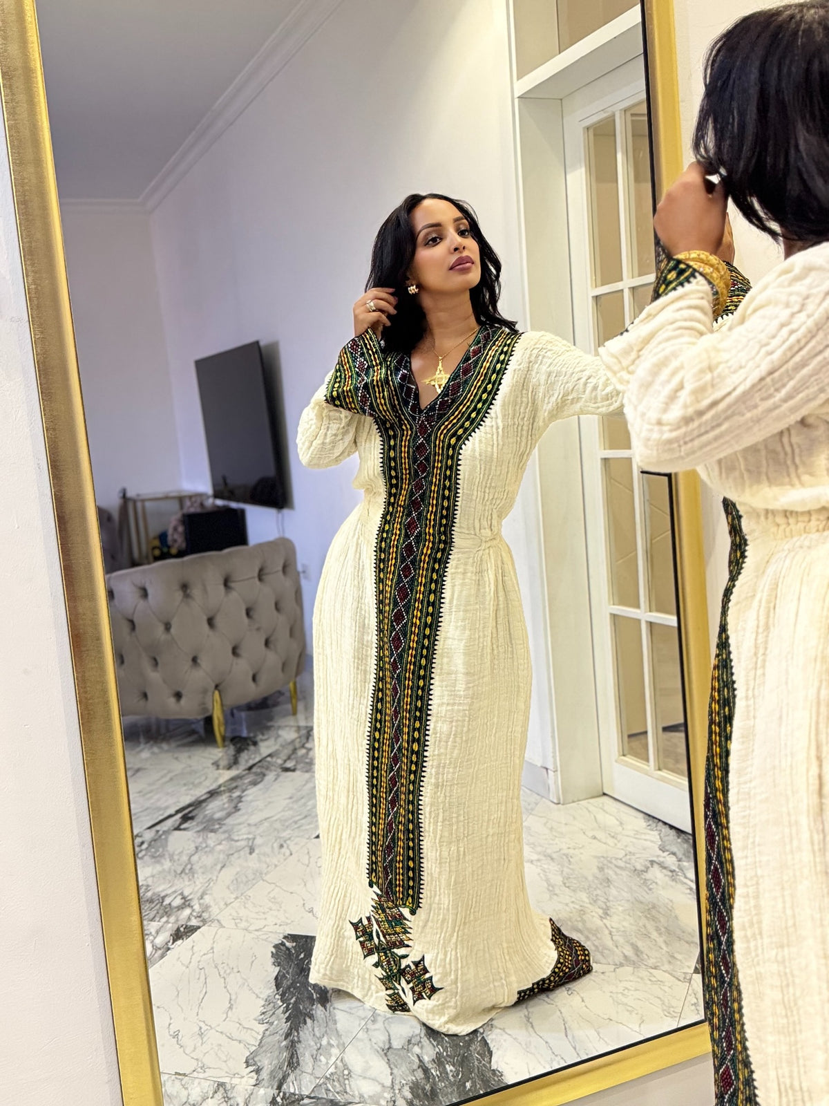 A woman wearing a long cream-colored Axum Fetil with green and yellow geometric embroidery, seen in a mirror reflection of a modern room with marble floors.