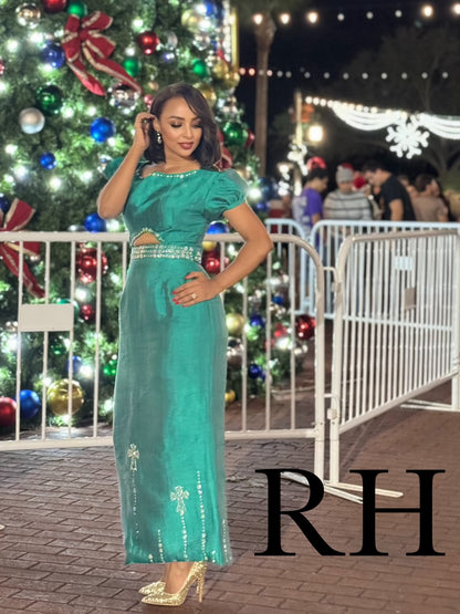 A woman modeling a teal green two-piece formal Ethiopian National Dress with gold embellishments, standing in front of a decorated Christmas tree at night. The background features festive lights, holiday decorations, and white crowd control barriers, with a large black 'RH' watermark overlay in the bottom right corner.