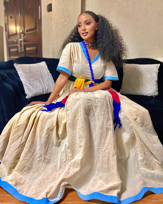 A woman wearing a voluminous cream-colored traditional Ethiopian National Dress with blue trim and colorful sashes sits on a dark couch in a home setting.