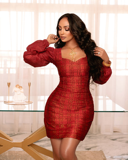 A woman modeling a red textured mini Axum Fetil with long puff sleeves and a sweetheart neckline, standing in a bright room with sheer curtains and a glass table.