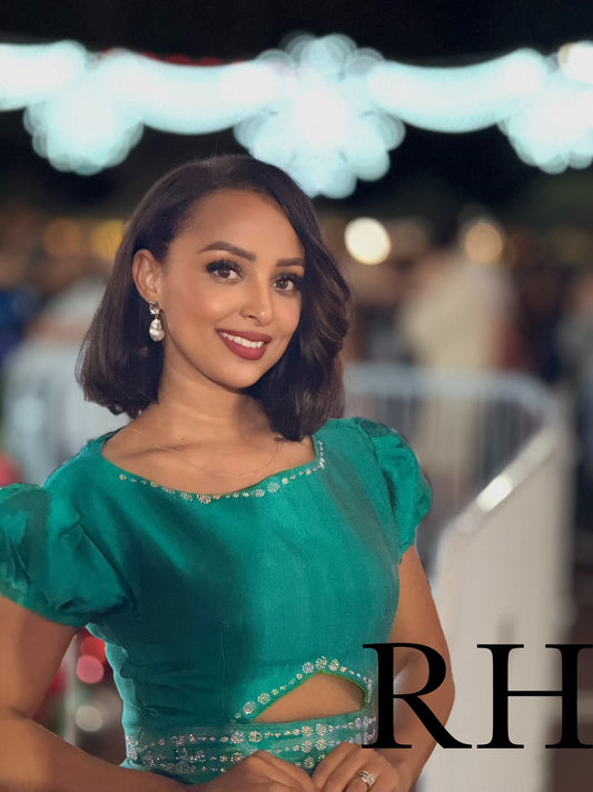 A woman with a short dark bob hairstyle wearing a vibrant emerald green traditional-style Ethiopian National Dress with puff sleeves and a cutout waist, posed against a blurred night event background with lights.