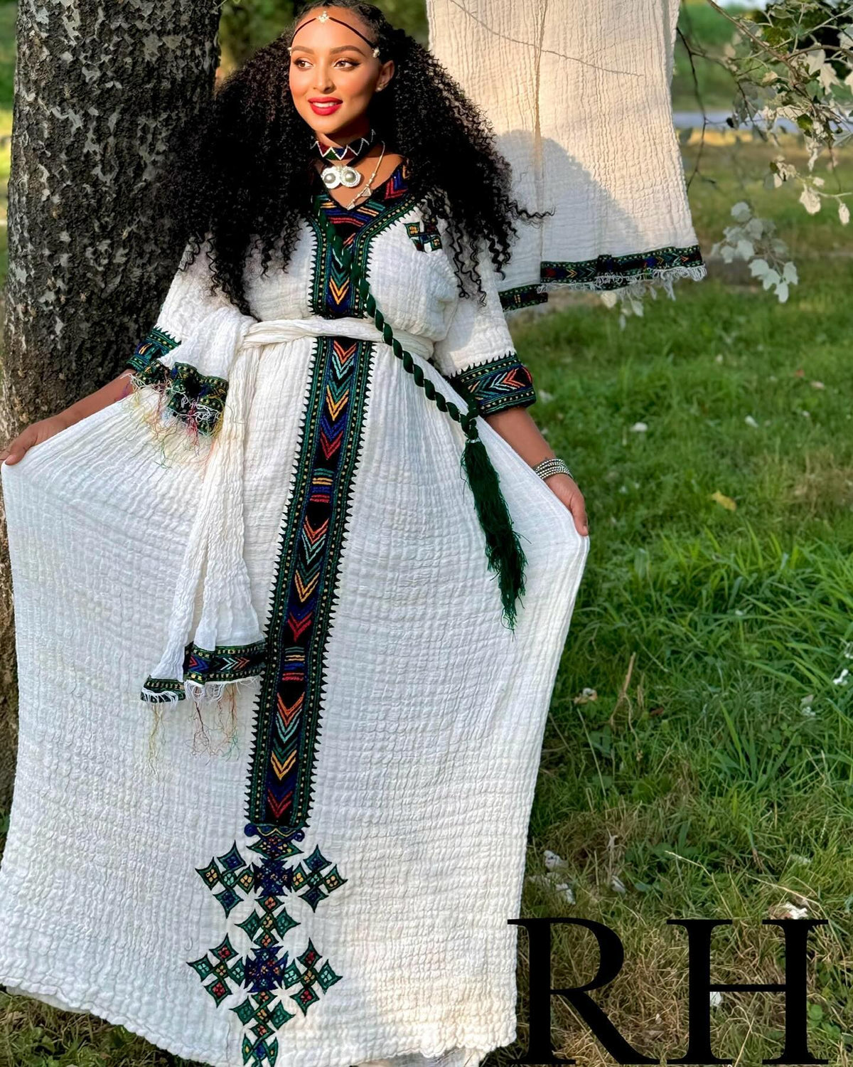 A woman wearing a long white textured Ethiopian National Dress with colorful geometric embroidery and a matching belt, standing outdoors next to a tree with grass in the background.