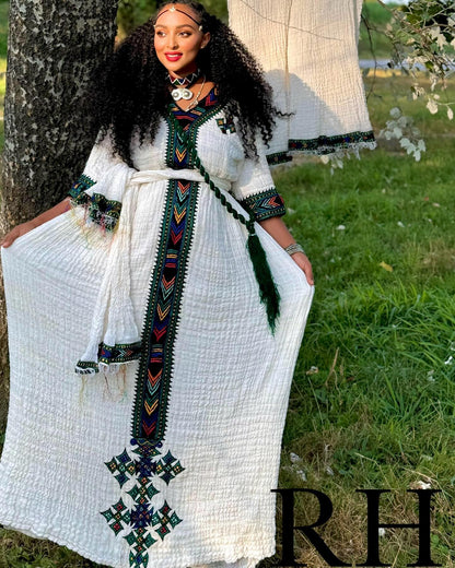 A woman wearing a long white textured Ethiopian National Dress with colorful geometric embroidery and a matching belt, standing outdoors next to a tree with grass in the background.