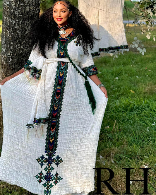 A woman wearing a long white textured Ethiopian National Dress with colorful geometric embroidery and a matching belt, standing outdoors next to a tree with grass in the background.