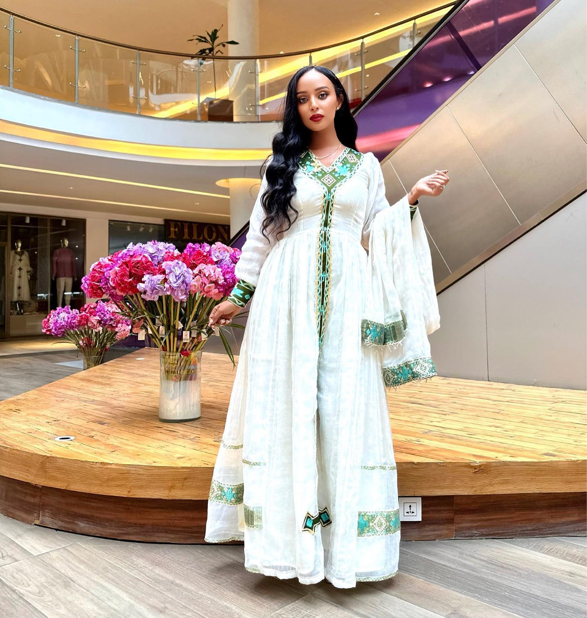 A woman models a traditional white long Ethiopian National Dress with green and gold embroidery in a modern indoor shopping mall setting with escalators and flower arrangements.
