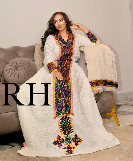 A woman models a long white  Axum Fetil with vibrant multicolor geometric embroidery and a matching embroidered shawl draped over a chair in a living room setting. A large 'RH' watermark is visible on the left side.