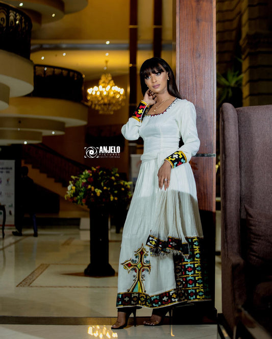 A woman wearing a long white traditional Axum Fetil with colorful embroidered details stands in a hotel lobby. The dress features a V-neck, long sleeves, and a wide hem decorated with a large cross motif and vibrant multicolor patterns.