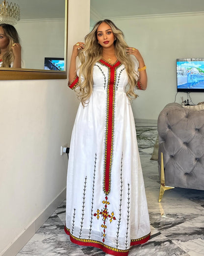 A woman wearing a long white embroidered Ethiopian National Dress with red and green detailing, standing in a modern living room with a gray tufted sofa and a mirror behind her.