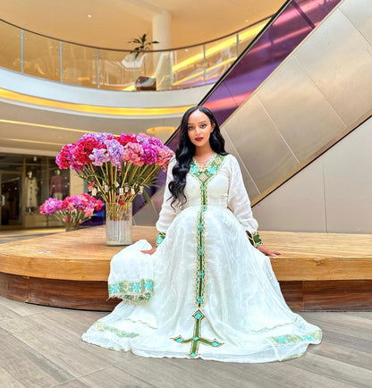 A woman wearing a traditional white embroidered Ethiopian National Dress with green and turquoise accents, posing in a modern multi-level indoor mall setting with escalators and flower arrangements in the background.