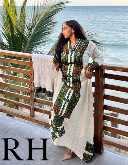 A woman stands on a beach balcony wearing a long white embroidered Axum Fetil with green, red, and gold detailing, holding a matching white throw blanket. Palm leaves and the ocean are visible in the background.
